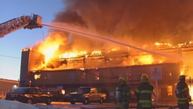 A large firetruck with a hose and ladder shoots water on a huge fire in an industrial building. A few firefighters look on.