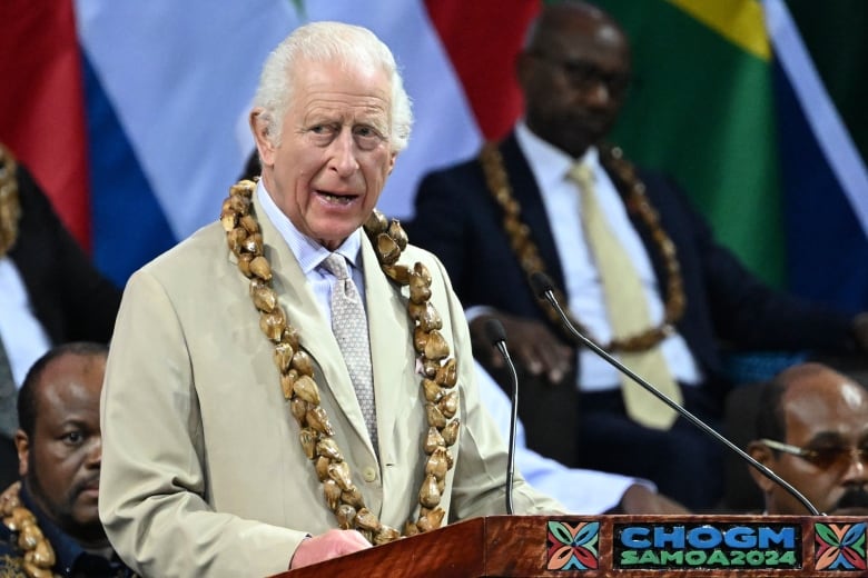 An older cleanshaven man wearing a light coloured suit with tie and a garland over top of it, speaks at a podium.