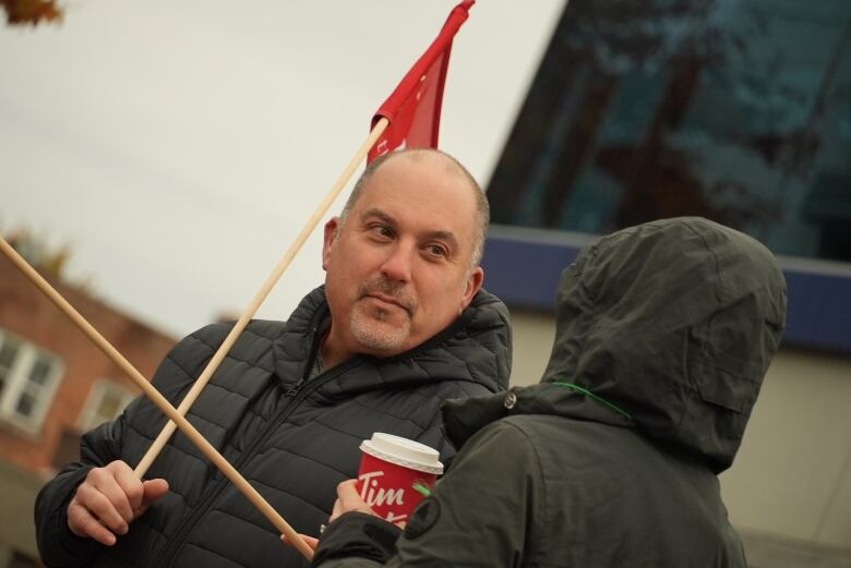 A man holds a red Unifor flag next to someone wearing a rain parka and holding a Tim Hortons cup