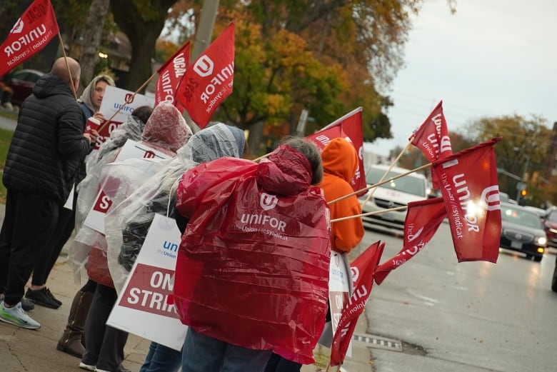 A group of people wearing red rain coats with the Unifor logo stand on the sidewalk in drizzling rain on a grey morning