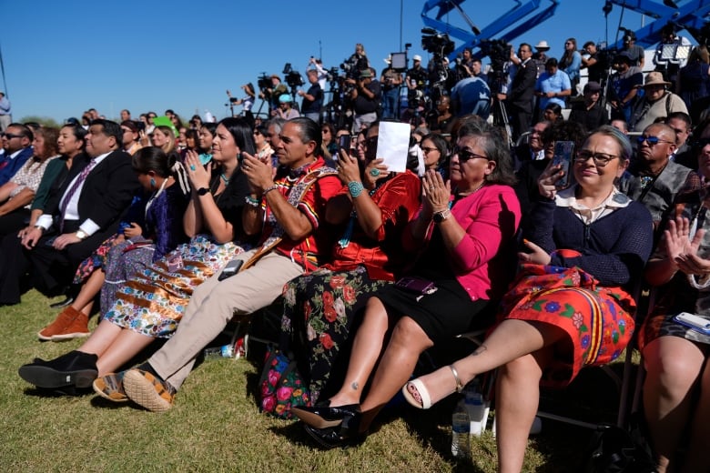 A crowd of people listens to a speech from a politician on a bright sunny day. Several people are clapping.