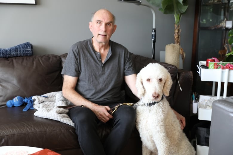 A man faces the camera and sits on a brown leather sofa with a large white curly-haired dog seated at his left.