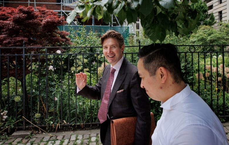 A lawyer in a suit and red tie smiles and waves to the camera.