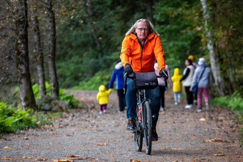 A man in an orange jacket riding a bike on a trail with people walking in the background. 