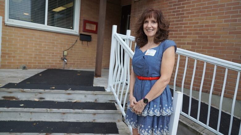 A woman in a light blue dress stands on the steps of a brick building. She's grinning at the camera.