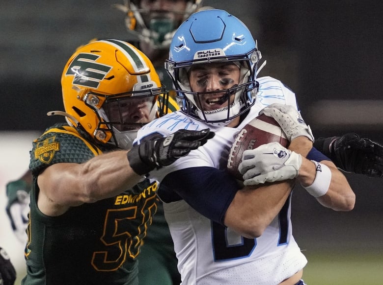 A football player in a white-and-blue uniform is running, cradling the ball. He is getting hauled down by an opposing player in a green-and-yellow uniform.