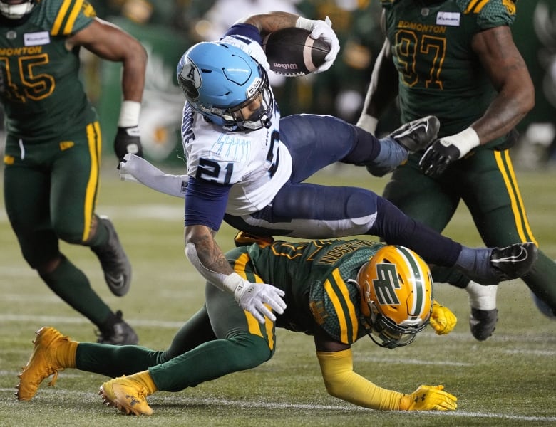 A football player in a white-and-blue uniform, holding the football in his left hand, is midair after an opposing player swept his legs out from under him.