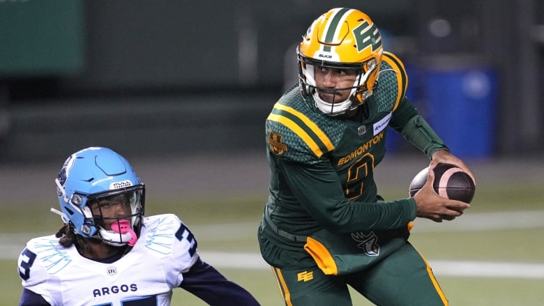 A football player in a green-and-yellow uniform is running on the field with the football in his hands. An opponent, in a white-and-blue uniform, is sitting on the field, watching.