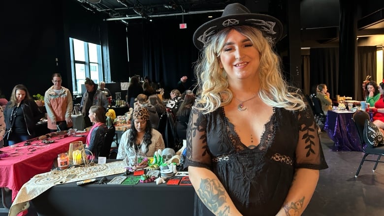 A woman dressed in a black witch's hat and dress standing in the foreground of a room filled with vendors wearing various witch costumes. 