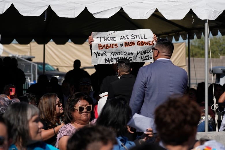 Someone standing up holding a sign that says 'There are still babies in mass graves. Your apology means nothing' behind a seated crowd, in front of two people facing toward them.