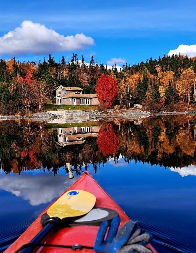 A kayak on a still body of water with a home next to trees with green, red and yellow leaves.