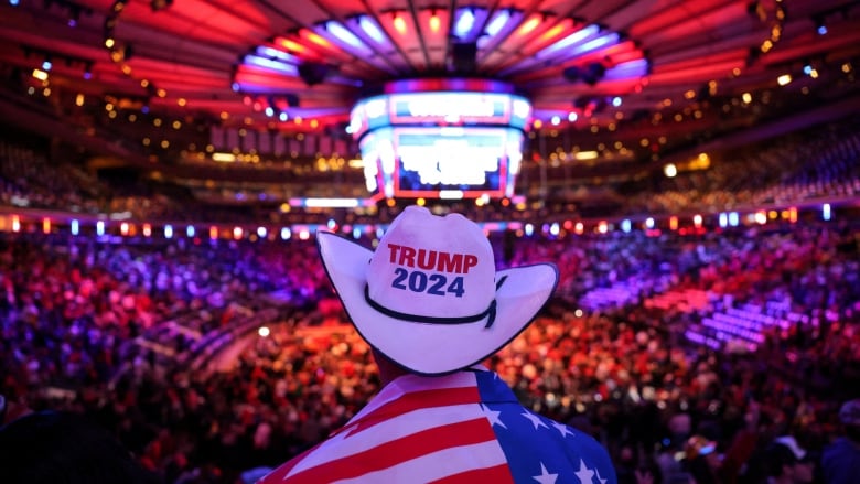 A person draped in a U.S. flag wears a cowboy hat that reads, 'Trump 2024,' inside a packed stadium.