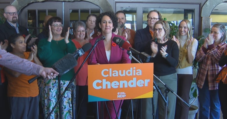 A woman with dark hair in a purple suit stands at a podium surrounded by cheering people