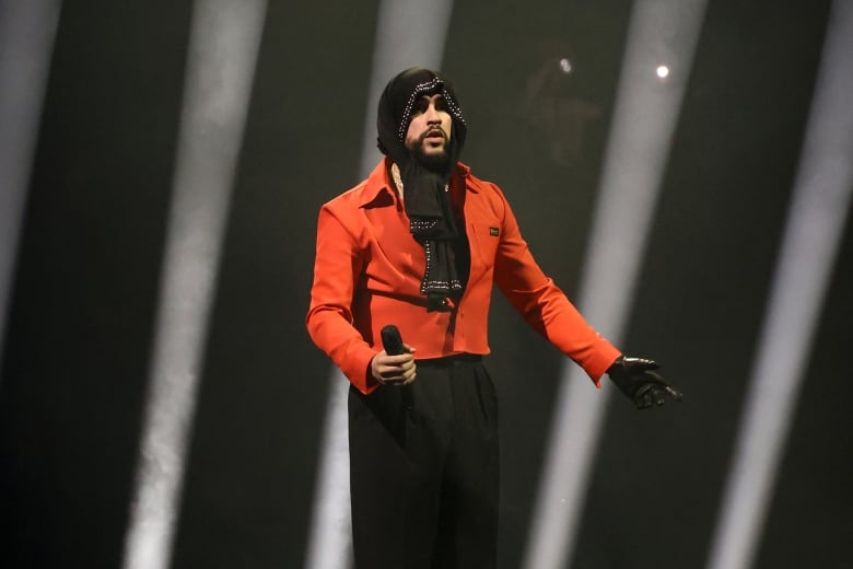 A bearded man wearing a hood and a glove on his left hand is shown holding a microphone while standing on a stage.