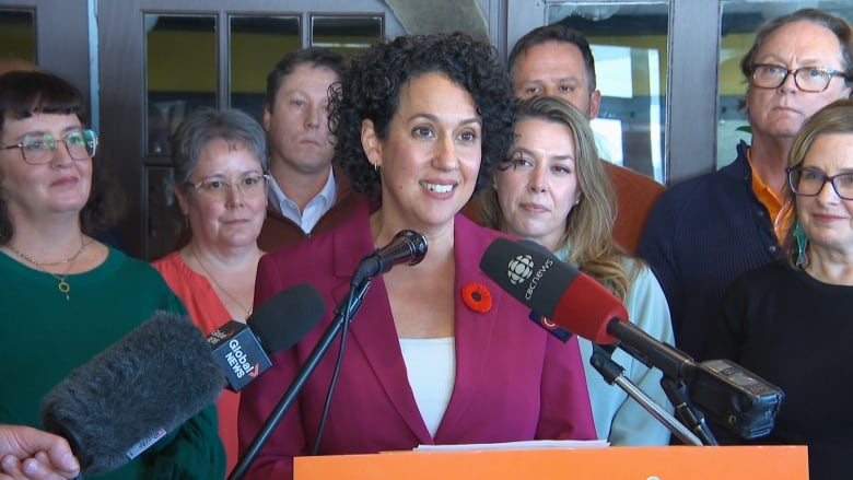 A woman in a pink suit speaks at a podium, surrounded by people.