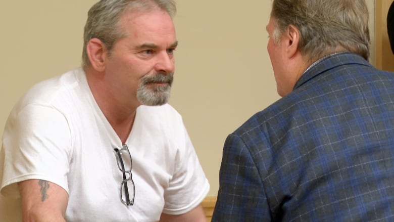 An older man in a white T-shirt standing facing another man in a blue and black suit. 