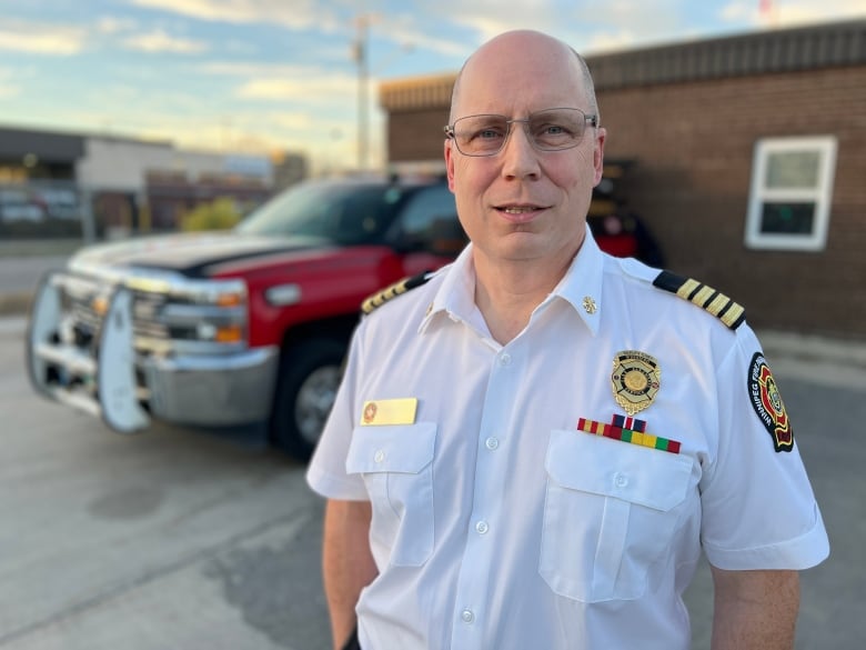 Man with glasses stands outside a firehall wearing a white uniform style shirt. 