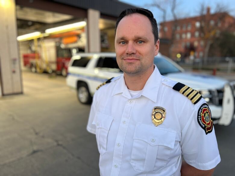 Man with dark hair stands outside a fire hall. He is wearing a white uniform style shirt. 