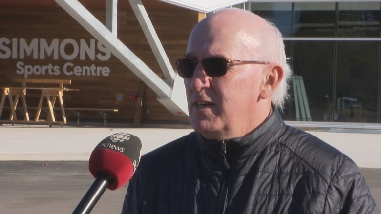 A man with sunglasses wearing a black jacket standing in the parking lot of an arena with a sign reading Simmons Sports Centre.