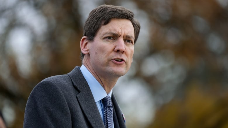 A white man looks to his right and speaks in front of a blurred background.