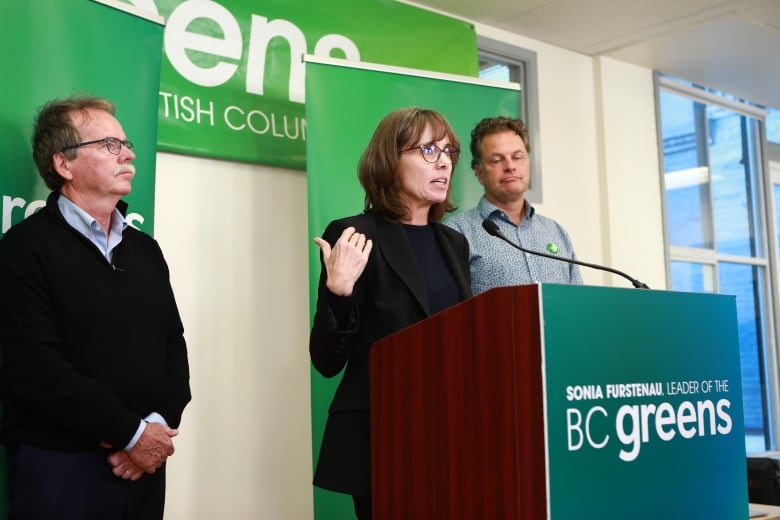 A woman answers questions at a podium, flanked by two men.