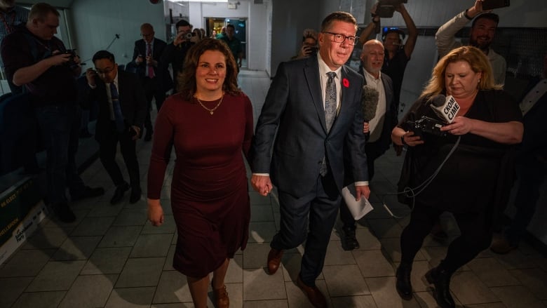 A man and woman walk down a hallway holding hands.