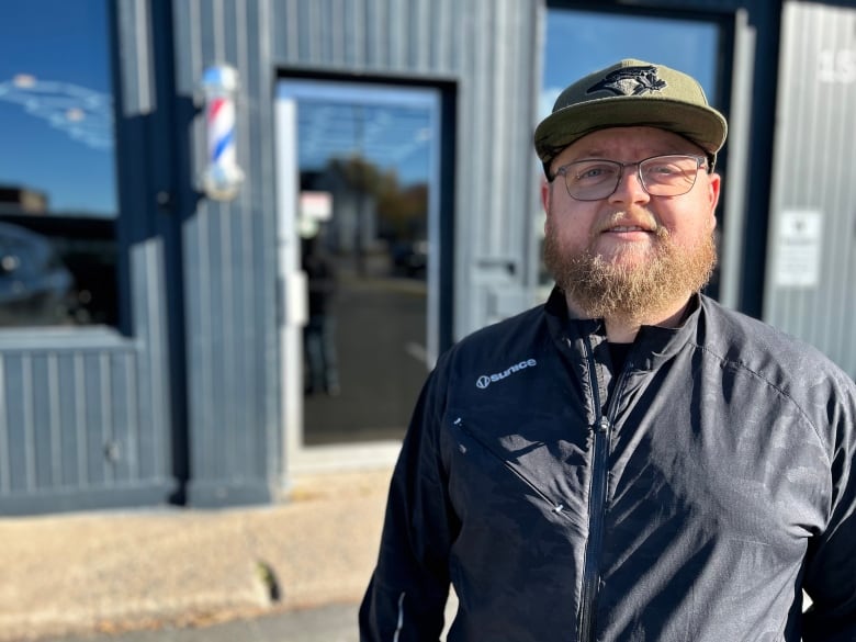 A man with glasses, a beard and a ball cap smiles at the camera in front of a black barbershop.