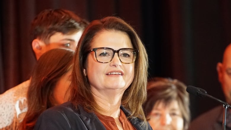 A woman wearing a grey suit jacket and orange shirt stands at a podium with people behind her.