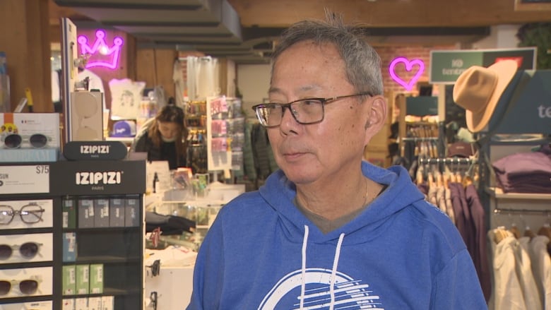 A man in a blue hoodie is interviewed in a gift shop with its wares displayed behind him.