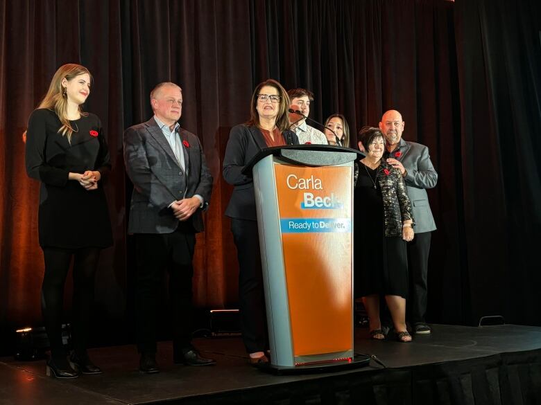 A woman stands in from of an orange podium surrounded by family members.