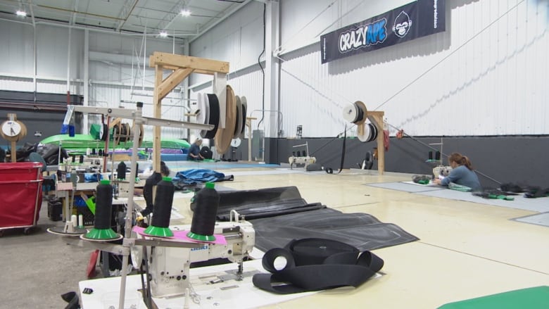 A distant angle of a warehouse that has a long table with sewing machines lined up the table and a woman is working at the far right of the table. 