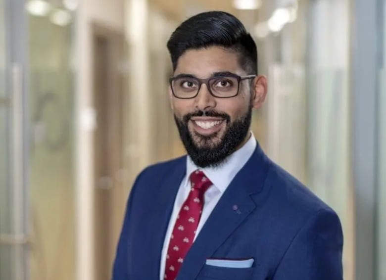 A man wearing black glasses and a suit jacket with a red tie smiles at the camera.