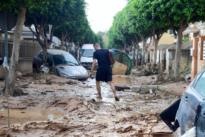 A short haired person with their back to camera wearing shorts tries to navigate a muddy street, with several damaged vehicles shown in the photo.