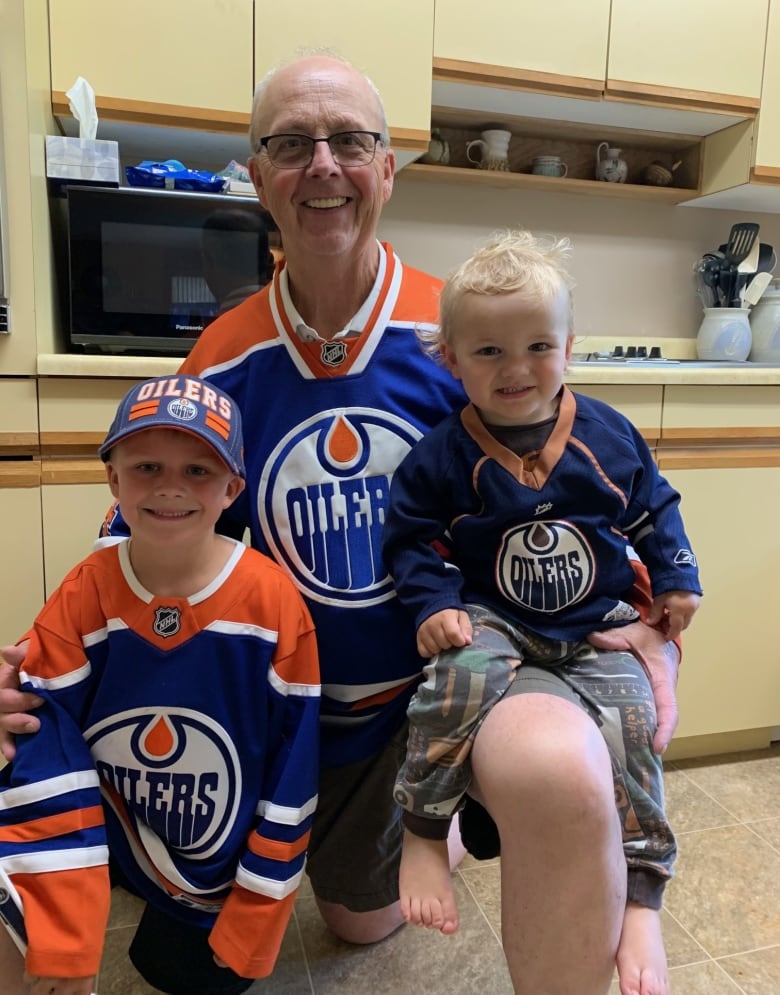 A man kneels next to two young children for a photo. They are all wearing Edmonton Oilers jerseys.