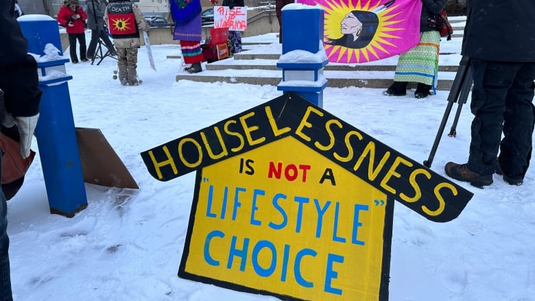 People stand outside a building. There is snow on the ground. Homemade signs and banners read 