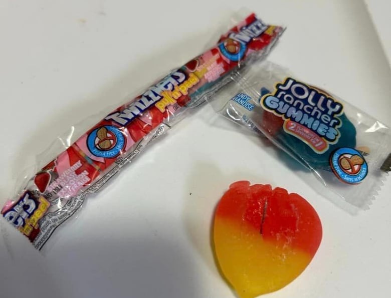 A package of Twizzlers licorice, a packaged Jolly Rancher gummy and an unpackaged gummy sit on a white table. The unpackaged candy has what appears to be a piece of metal poking out of the centre.