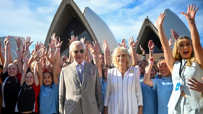 Two people stand in front of a crowd of people with their hands raised in front of the Sydney Opera House.