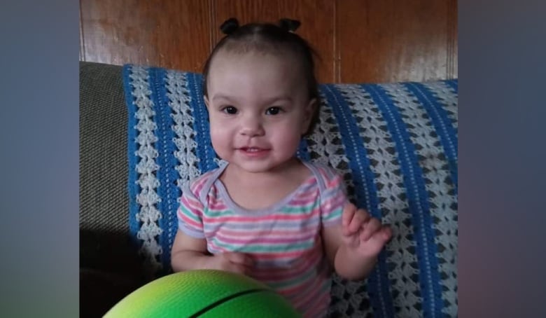 An infant girl with hair in space buns sits on a couch and smiles, with a basketball on her lap.