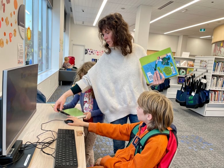 A woman with long dark hair helps a young child sitting at a library desk with a computer.