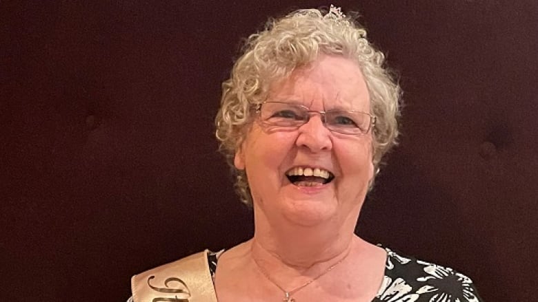 A senior woman with short, curly white hair and rectangular glasses smiles.