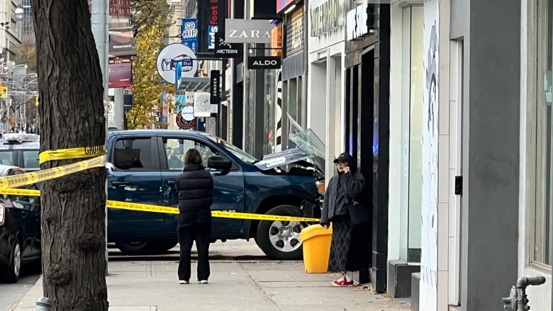 A truck is crashed into a city storefront. Two people look on. It's a grey day. Glass is broken on the sidewalk. Police tape is wrapped around the scene.