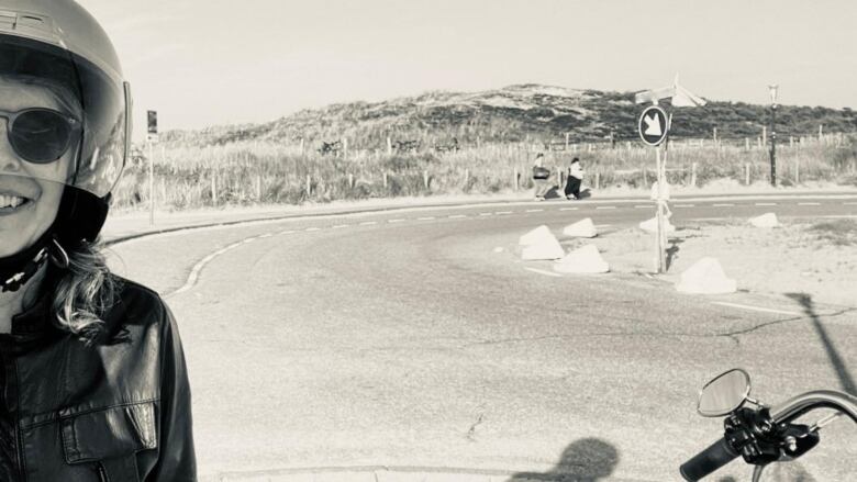 A woman with a black motorcycle helmet and leather jacket in the corner of a sepia photo of a road