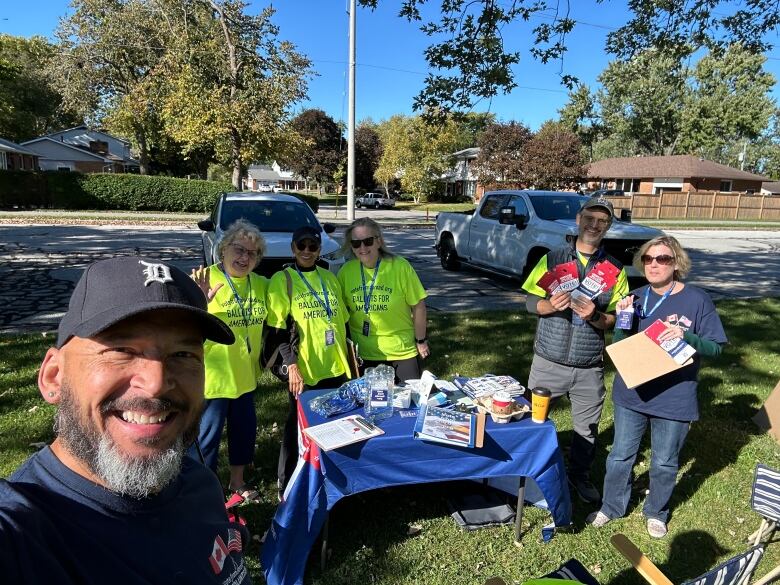 A group of people take a selfie gathered around a table outdoors. Some of them are holding pamphlets, wearing lanyards and neon yellow shirts.