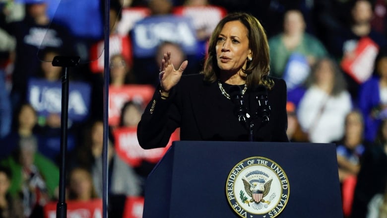 Kamala Harris, wearing a black suit with her signature necklace, raises her arm as she speaks at a podium in front of a crowd of supporters. 