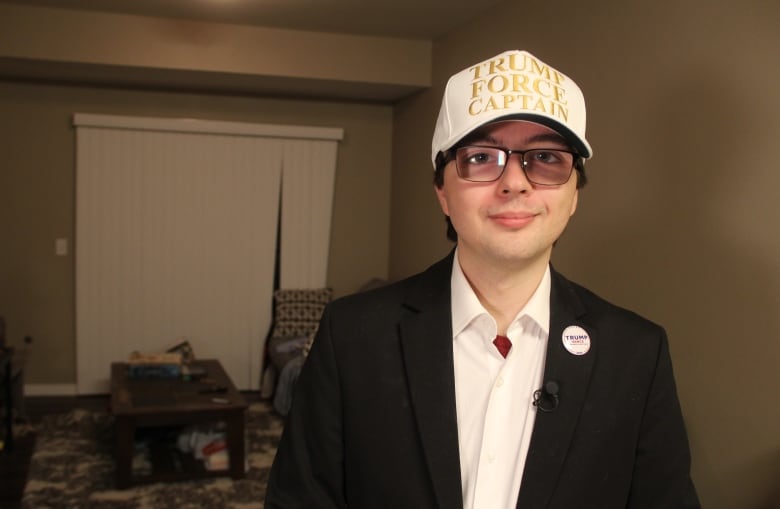 A man in a black blazer and white buttoned-up shirt underneath stands in a living room, while wearing a white hat with the letters in gold reading, 