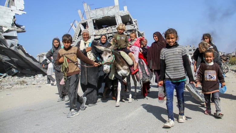 A group of women and children walk through a street next to buildings that have been reduced to rubble.