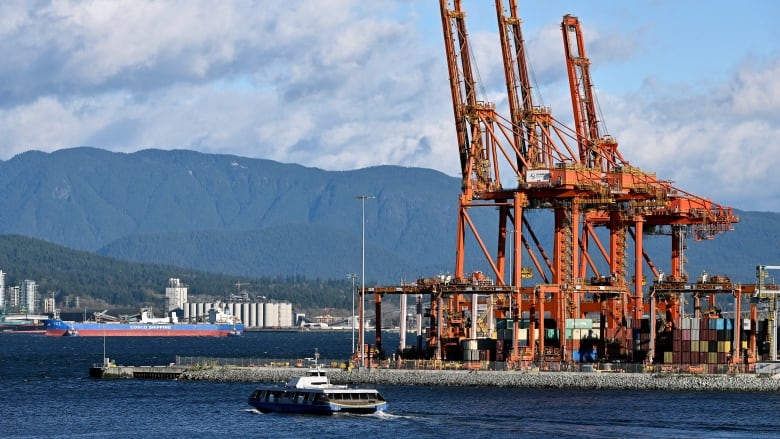 Big orange cranes are seen at the right side of the image of a port. There are mountains in the background and a small boat passing in front of the port in the water. A big container ship is seen in the distance. 