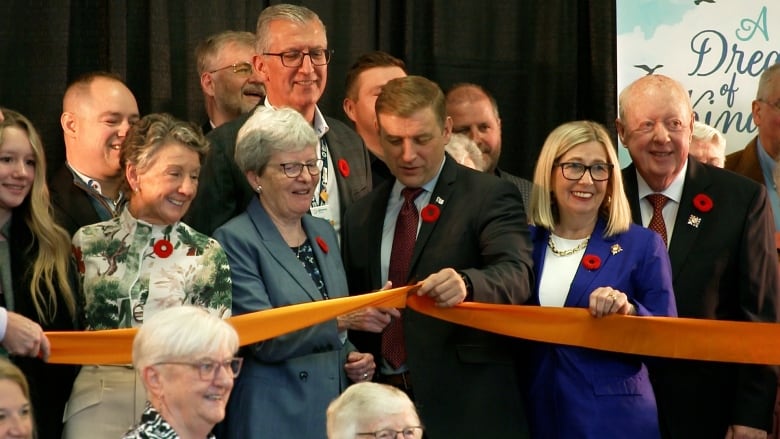 A group of people crowd around a long orange ribbon being cut by a woman standing in the centre.