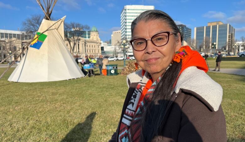 A woman wearing a brown jacket and black glasses smiles to the camera, with a teepee behind her.