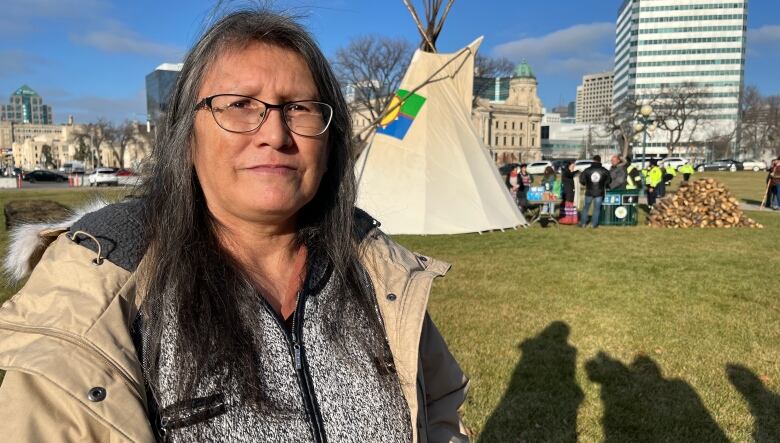 A woman with medium-length hair, wearing a jacket and sweater, looks to the camera, with a teepee behind her.